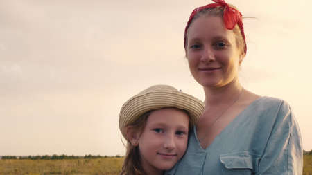 Portrait of a beautiful mother with her daughter who are standing in an embrace on a beautiful field at sunset. Blonde mother and daughter with hats.の写真素材