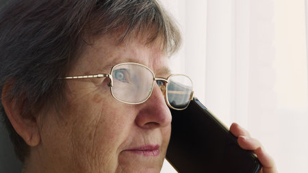 An elderly woman with glasses stands by the window and talks on the phone with her loved ones. An...