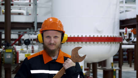 An oil and gas worker stands in a hard hat and ear protectors in a gas liquefaction shop, behind him is a container of gas, and in his hands is a large wrench. Workplace repairman.の写真素材