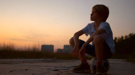 A teenage boy in the evening in the city sits on a ball and dreams of the future sports career of a football player, future soccer celebrity. The childhood of a great soccer player.の写真素材