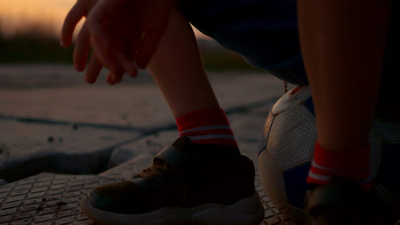 Close-up of a guy sitting on a soccer ball. A teenager in sneakers sits on a ball resting after the game, in the rays of the setting sun. Youth and boyish ball games.の写真素材