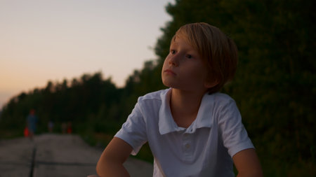 Close-up of a blond guy sitting in a park at sunset. A portrait of a teenager in a white T-shirt who sits and enjoys the setting sun. Youth and boyish dreams.の写真素材