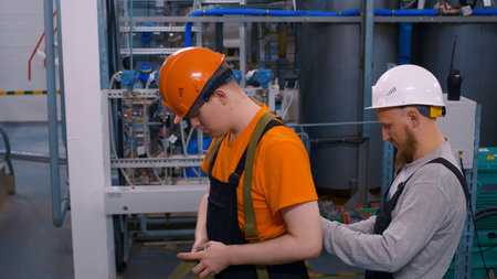 Two men in hard hats at work in a chemical plant. One worker puts on a work belt, his senior foreman...