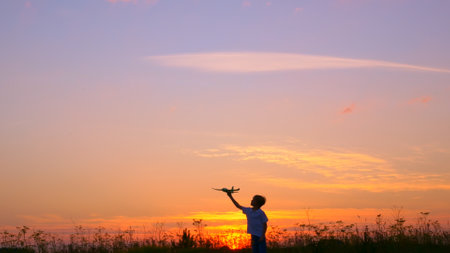A teenage boy runs through the meadow in the rays of the setting sun. The boy has a toy plane in his hands. The boy dreams of flying on an airplane like a bird. Childrens dreams of the futureの写真素材