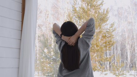 A woman sits on the edge of the bed, looks out the window and stretches after sleep, outside the...