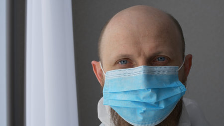 Close-up of a doctors face in a surgical mask. A bearded medic takes off his mask after working with...