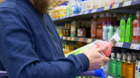 The buyer in the store chooses shampoo on the shelves. Buying personal care products for men, buying liquid soap in the supermarket. A man chooses a product carefully reading the composition.の写真素材