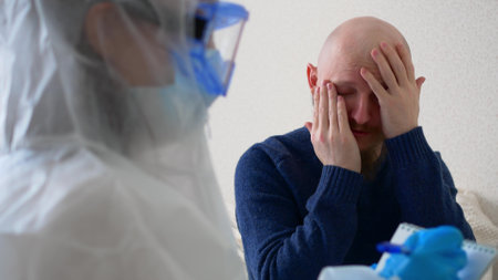 A sick man complains to the doctor about his health, the Man coughs and says that his throat hurts. A doctor in a protective suit, mask and goggles writes down the symptoms of the disease.の写真素材