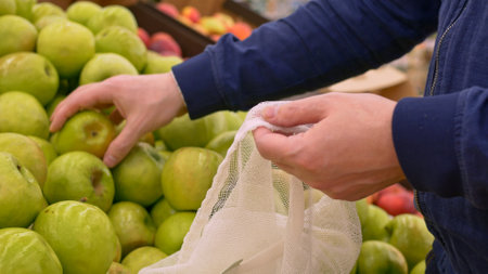 Buying green apples in the supermarket. A juicy apple is taken from the farmers market shelf and put into a reusable bag. Eco fruits for vitamins. Using reusable shopping bags in the store.の写真素材