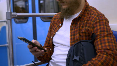 A man with a beard sits in a subway car and has fun on the Internet using a smartphone. Pass the...
