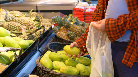 A man in a supermarket in the fruit section chooses the most delicious and ripe mangoes and puts them in a white string bag. Following a proper and healthy diet. Concept of buying fruits by a man.の写真素材