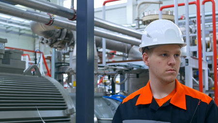A young engineer in a white helmet and overalls at his workplace looks at the drawing of the...
