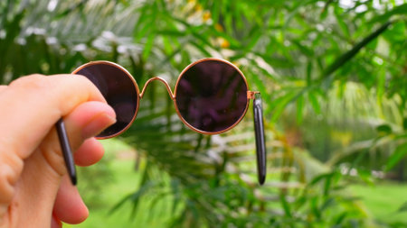 Sun glasses are held by unrecognizable female hands against backdrop of picturesque rainforest. Hands hold oval round glasses. Beautiful accessory, round sunglasses. Blurred rainforest in backgroundの写真素材