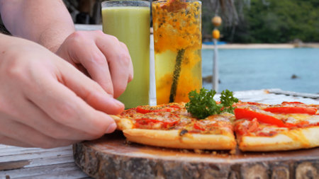 Delicious food in a restaurant by the sea. Cheesy pizza and raw vegan drinks for lunch. Concept of a healthy vacation with delicious food on vacation. Close up hand takes delicious food cheesy pizza.の写真素材
