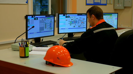 Focused worker in industrial setting scrutinizes equipment performance on monitor. Worker operates...