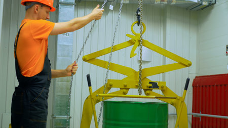Worker lifts chemical waste barrel with chain hoist in industrial plant. Emphasizing safety protocols, protective gear. Chemical waste handling requires precision, caution. Worker ensures secure.の写真素材