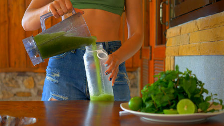 Woman pouring refreshing green smoothie into glass with ice, accompanied by fresh mint and lime slices on table. Woman is pouring green smoothie from bottle into glass with ice. Healthy eatingの写真素材