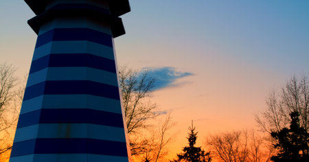 Decorative blue and white striped lighthouse isolated on white background, set against a backdrop of a beautiful sunset with orange, pink, and blue hues blending seamlesslyの写真素材