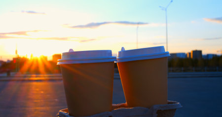 Sunlight shines between two disposable coffee cups on a cardboard tray, creating a starburst effect, with a blurred urban background at sunsetの写真素材