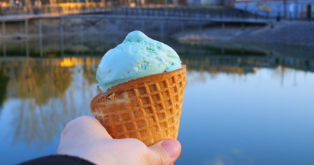 Hand holding creamy ice cream cone near tranquil lake surface, reflecting warm sunset colors with...