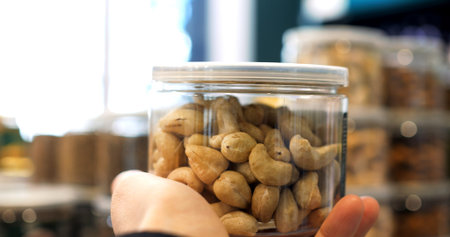 Hands holding a transparent plastic jar filled with cashew nuts, inside a grocery store, with...