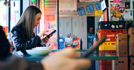 Young woman casually dining in vibrant restaurant, browsing smartphone while enjoying meal,...