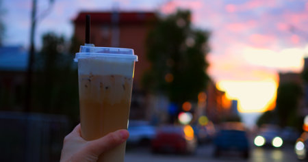 Hand gripping chilled coffee drink with whipped cream, straw visible against blurred urban sunset, background featuring traffic lights, city buildingsの写真素材