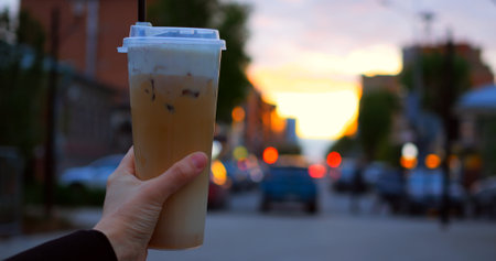 Female hand holding frothy cold coffee beverage, standing amid urban street during golden hour, blurred cityscape creating soft luminous backdropの写真素材