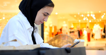Young adult muslim woman wearing hijab drinking coffee and using smartphone while sitting in shopping mall food court, enjoying leisure moment with modern technology and relaxed urban lifestyleの写真素材