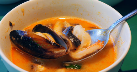 Savory tom yum soup brimming with succulent seafood, mussels, shrimp, squid, and mushrooms, carefully lifting from creamy white bowl with metal spoonの写真素材
