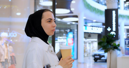 Young muslim businesswoman in hijab enjoying coffee break, working on tablet while walking through modern shopping mall interiorの写真素材