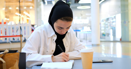 Focused muslim student girl in hijab is making notes in notebook, sitting in cafe at shopping mall food court, studying and drinking coffee.の写真素材