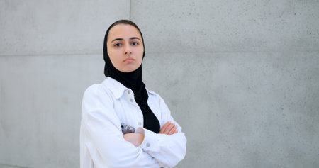 Serious young muslim professional wearing hijab and white shirt, confidently standing against gray wall with arms crossed and direct gazeの写真素材
