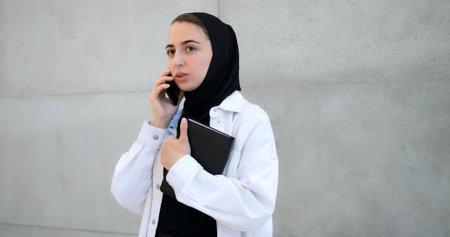 Professional businesswoman wearing hijab standing near gray wall while making phone call, holding notebook with focused expression during work communicationの写真素材