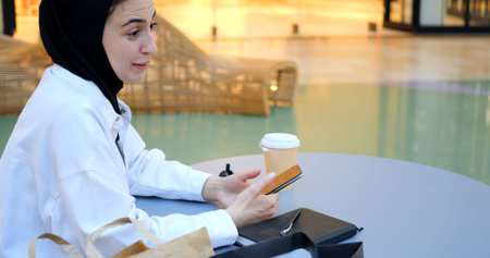 Young muslim woman wearing traditional hijab sitting at a table in a shopping mall, using her...