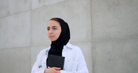 Young muslim woman in hijab leaning against concrete wall, holding book and gazing thoughtfully, reflecting on academic goals and professional aspirationsの写真素材