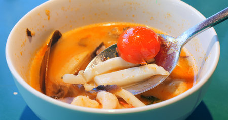 Metal spoon lifting mussels and shrimp from white bowl, revealing vibrant ingredients of traditional tom yum soup highlighting rich thai culinary flavorsの写真素材