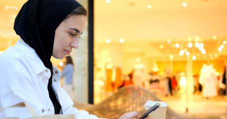 Young muslim woman wearing a hijab enjoys a coffee while using her smartphone in a brightly lit shopping mall, showcasing a blend of modern life and tradition. Free Islamic woman in a shopping centerの写真素材