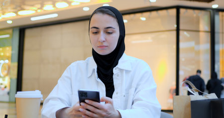 Young entrepreneur wearing hijab reviews notes and uses tablet in bustling shopping center food court, showcasing modern work practices and cultural diversity. Muslim diligent student, flexibleの写真素材