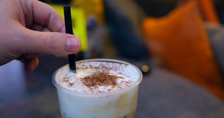 Female barista stirring frothy cappuccino, chocolate dust topping, relaxing near sunlit cafe window