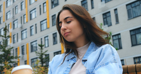 Stylish young woman sits on a bench, savoring a hot beverage from a disposable cup, taking a break amidst the backdrop of a modern buildingの写真素材