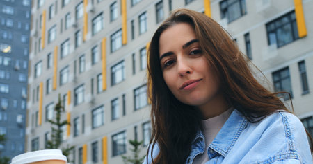 Attractive young woman with long brown hair, wearing a denim shirt, savors a takeaway coffee in front of a contemporary residential building, exuding a sense of urban tranquilityの写真素材