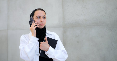 Professional businesswoman wearing hijab standing near gray wall while making phone call, holding notebook with focused expression during work communicationの写真素材