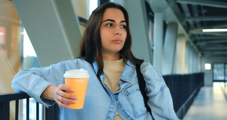 Student waiting impatiently, checking smartwatch time while drinking coffee from takeaway cup in bright, contemporary building corridorの写真素材