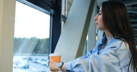 Young woman wearing denim jacket and smartwatch drinking hot beverage, gazing pensively through...