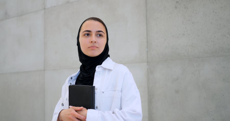 Muslim woman wearing hijab, white shirt, holding book close, gazing pensively near concrete wall, embodying contemplative momentの写真素材
