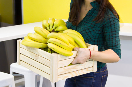 Female hands hold a wooden box with bananas in the kitchen. Banana on wooden basketの写真素材