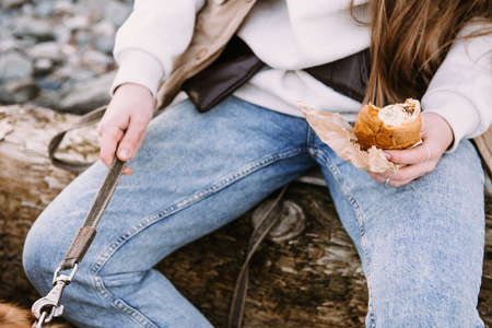 young woman walking with dog on river shore and eating croissantの写真素材