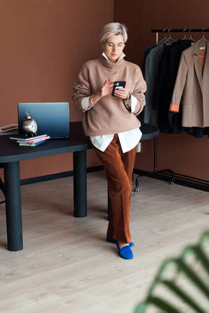 fashion woman in brown oversize sweater and trousers standing in modern work place or office with smartphoneの写真素材