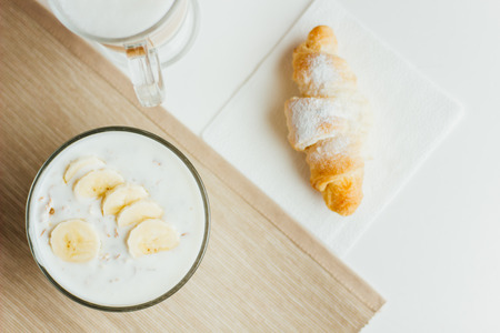 Yogurt with muesli and banana in a transparent bowl, croissant and coffee on a gray napkinの写真素材